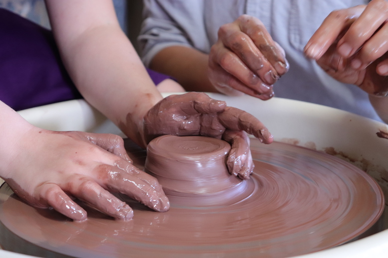Kids hands using red clay on a potter's wheel with adult hands helping