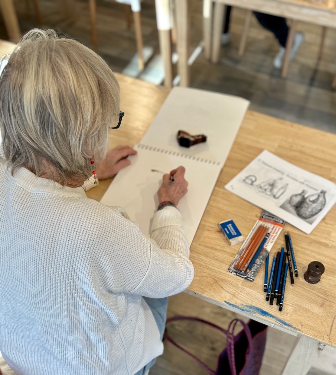 Over the shoulder of a woman sketching with drawing supplies on a table